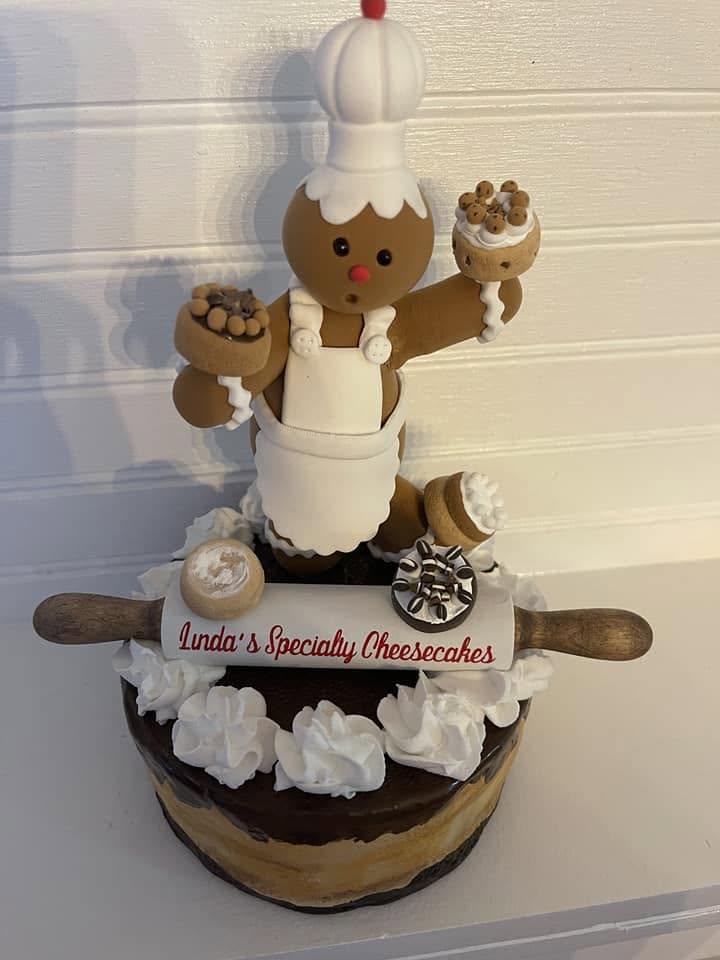 Linda's handmade clay gingerbread chef mascot standing on a cheesecake, holding the Linda's Specialty Cheesecakes rolling pin.