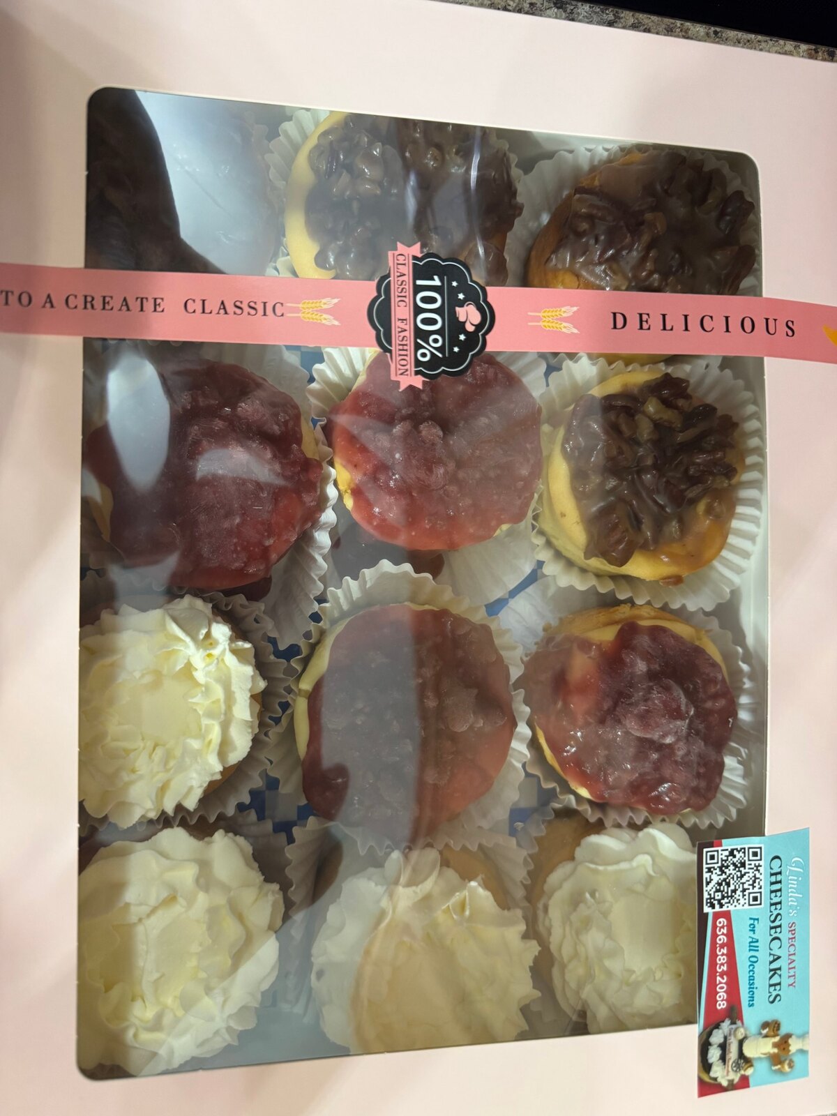 Windowed bakery box containing twelve assorted mini petite cheesecakes with fruit, cream, and chocolate toppings.