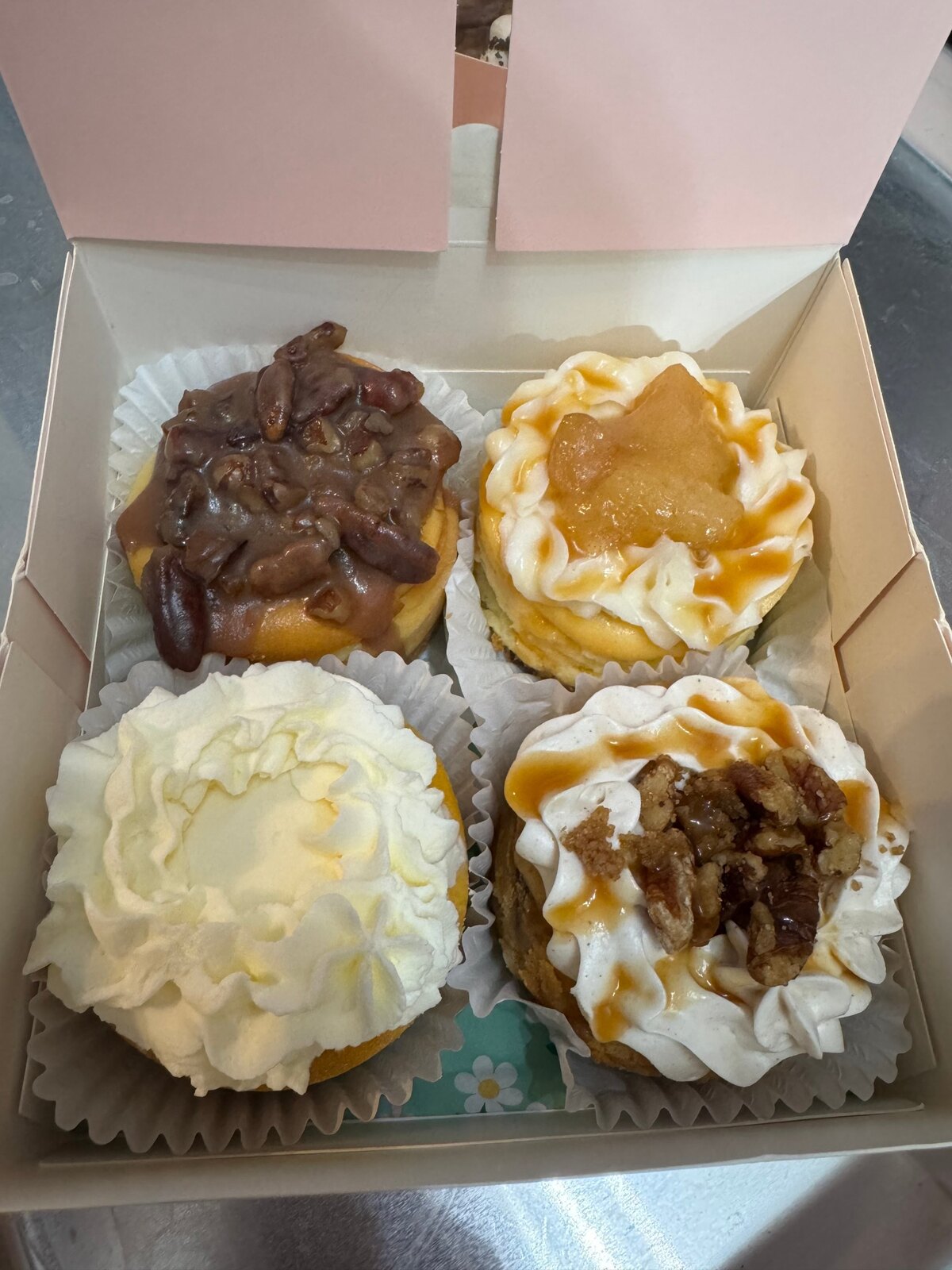 Open pink bakery box containing four mini petite cheesecakes with assorted fruit, cream, and pecan toppings.