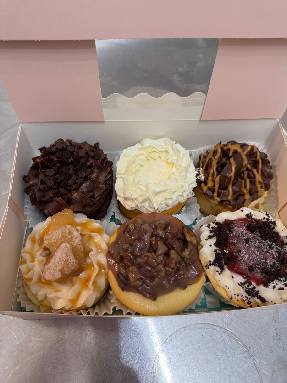 Open pink bakery box containing six mini petite cheesecakes with assorted chocolate, fruit, cream, and pecan toppings.